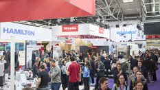 A crowd of people in an exhibition hall with various stands and visitors.