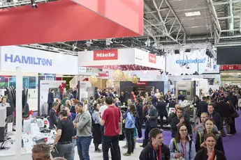 A crowd of people in an exhibition hall with various stands and visitors.