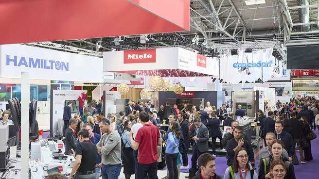 A crowd of people in an exhibition hall with various stands and visitors.