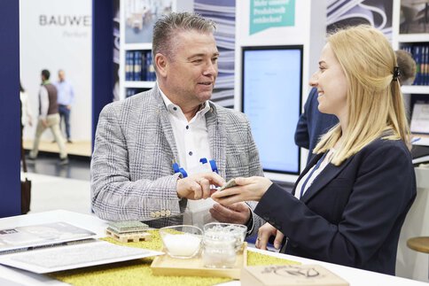 An einem Messestand der BAU hält eine Frau in Businesskleidung einem Mann in kariertem Jackett eine Materialprobe zum Befühlen entgegen.