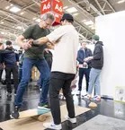 Ein Mann testet ein Balance-Board bei einem Messestand. Er bekommt Hilfe von einem weiteren Mann. Im Hintergrund sind weitere Messebesucher zu sehen.