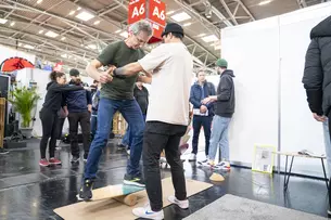 Ein Mann testet ein Balance-Board bei einem Messestand. Er bekommt Hilfe von einem weiteren Mann. Im Hintergrund sind weitere Messebesucher zu sehen.