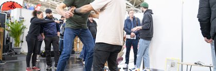 Ein Mann testet ein Balance-Board bei einem Messestand. Er bekommt Hilfe von einem weiteren Mann. Im Hintergrund sind weitere Messebesucher zu sehen.