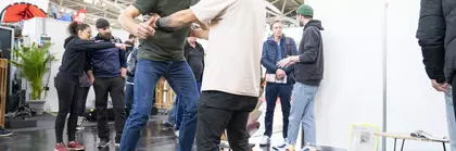Ein Mann testet ein Balance-Board bei einem Messestand. Er bekommt Hilfe von einem weiteren Mann. Im Hintergrund sind weitere Messebesucher zu sehen.