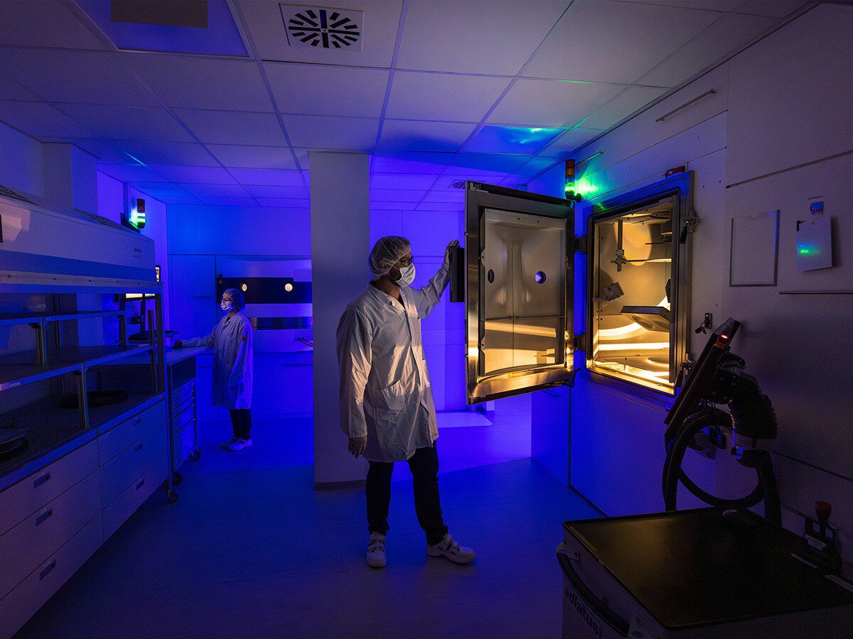 Two people in protective gear are working in a cleanroom laboratory illuminated by blue light and operating an open piece of equipment with an illuminated interior.