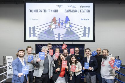 People with red and blue cards pose in a boxing ring. Above them, a screen announces the 