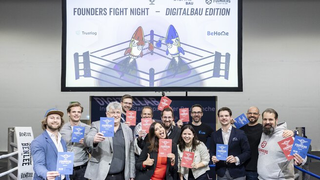 People with red and blue cards pose in a boxing ring. Above them, a screen announces the 