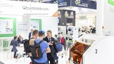Menschen unterhalten sich in einer hell erleuchteten Ausstellungshalle der productronica mit verschiedenen Ausstellerständen, Werbeplakaten und -bannern.