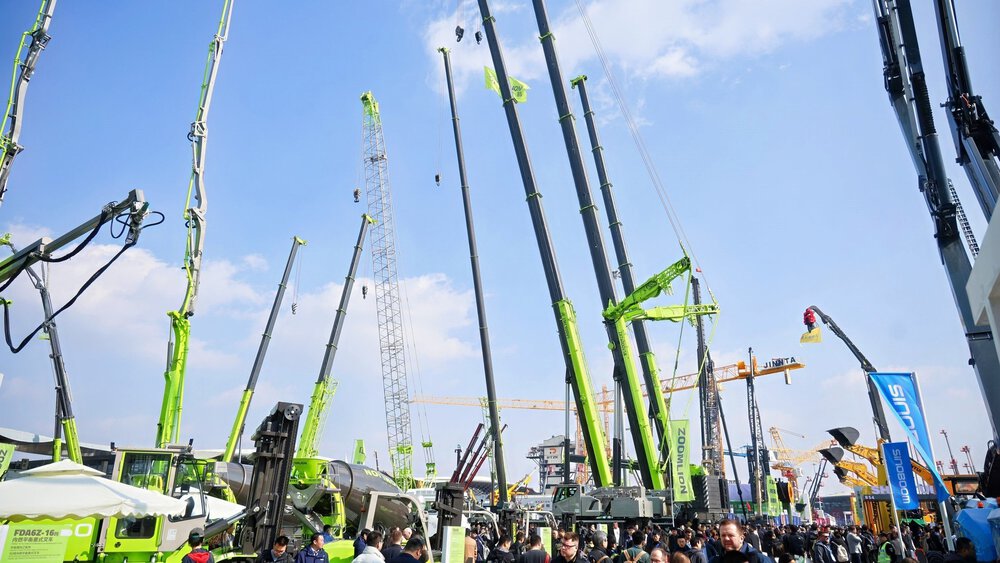 Under a blue sky, people stroll among large green construction cranes on display at an outdoor construction equipment exhibition.