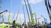 Under a blue sky, people stroll among large green construction cranes on display at an outdoor construction equipment exhibition.