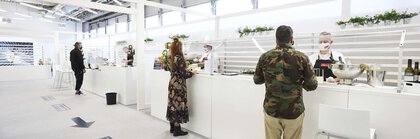 Menschen stehen an einer modernen weißen Essensausgabe, hinter der Köche mit FFP2-Masken Gerichte zubereiten.