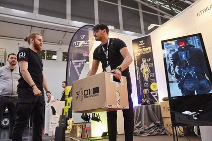 Ein Mann hebt unter der Aufsicht eines Ausstellers an einem Messestand einen schweren Umzugskarton an, mit Hilfe eines Exoskelets.