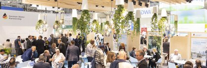 Ein gut besuchter Messestand mit sitzenden und stehenden Menschen, von der Decke hängenden Pflanzen und heller Beleuchtung.