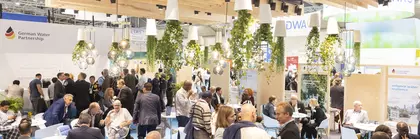 Ein gut besuchter Messestand mit sitzenden und stehenden Menschen, von der Decke hängenden Pflanzen und heller Beleuchtung.