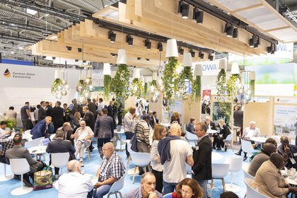 Ein gut besuchter Messestand mit sitzenden und stehenden Menschen, von der Decke hängenden Pflanzen und heller Beleuchtung.