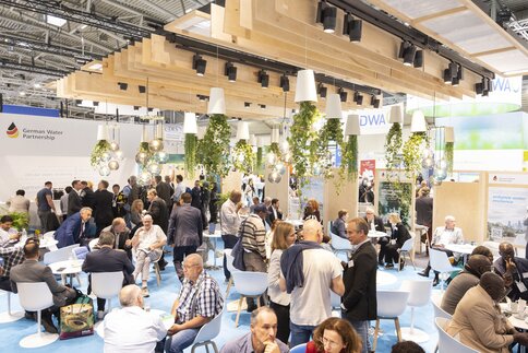 A well-attended exhibition stand with people sitting and standing, plants hanging from the ceiling and bright lighting.