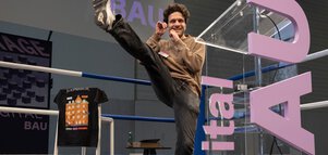 Auf der Startup-Bühne im Boxring-Look steht ein junger Mann mit gehobenen Fäusten und kickt mit dem rechten Bein in die Luft.