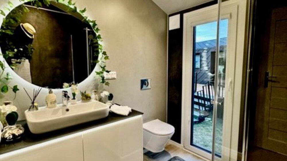 Modernes Badezimmer mit rundem Spiegel, Blätterdekor, weißem Waschbecken, Toilette und Glasdusche. Tür führt zur Terrasse.