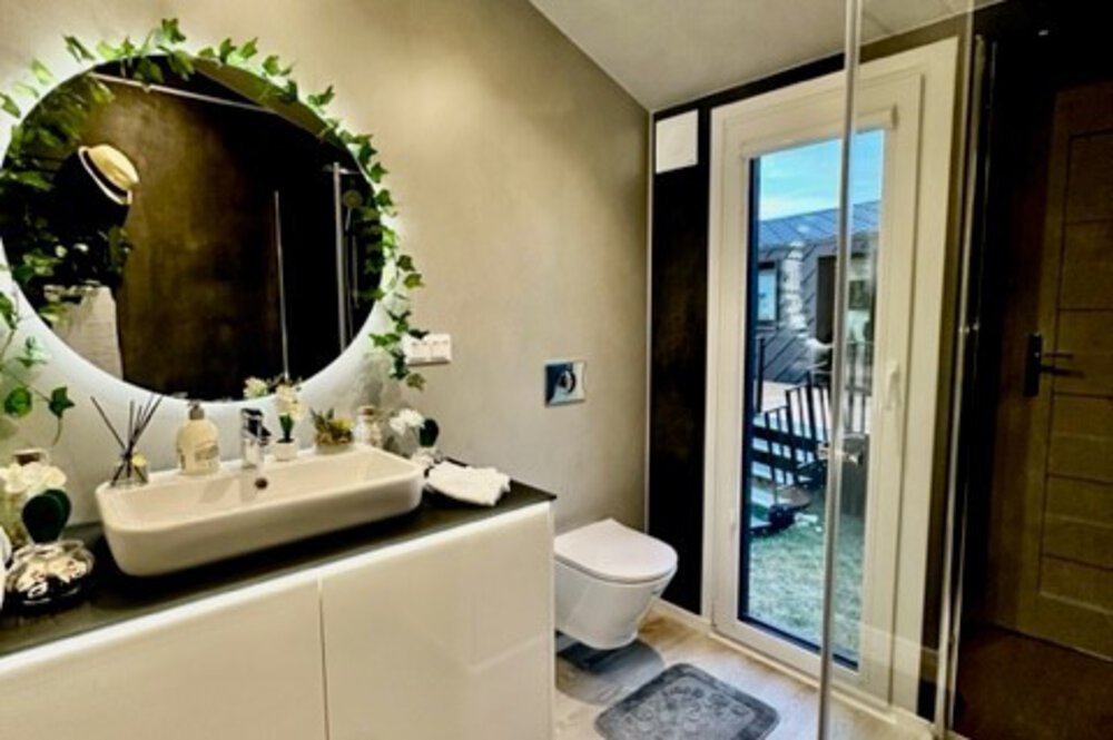 Modernes Badezimmer mit rundem Spiegel, Blätterdekor, weißem Waschbecken, Toilette und Glasdusche. Tür führt zur Terrasse.