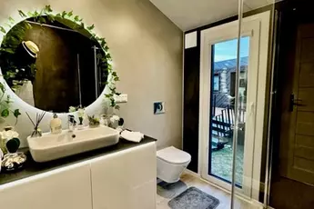 Modernes Badezimmer mit rundem Spiegel, Blätterdekor, weißem Waschbecken, Toilette und Glasdusche. Tür führt zur Terrasse.