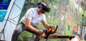 Mann mit VR-Brille simuliert Holzfällarbeit mit Motorsäge in einer interaktiven Messeinstallation vor Waldkulisse.
