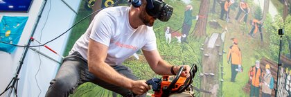 Man wearing a VR headset simulates woodcutting with a chainsaw in an interactive exhibition installation with a forest backdrop.