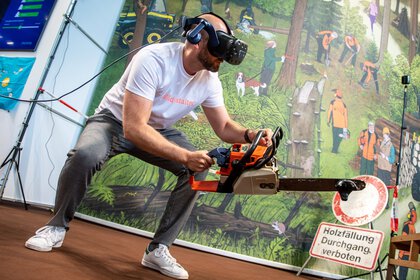 Mann mit VR-Brille simuliert Holzfällarbeit mit Motorsäge in einer interaktiven Messeinstallation vor Waldkulisse.