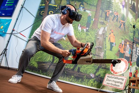 Man wearing a VR headset simulates woodcutting with a chainsaw in an interactive exhibition installation with a forest backdrop.