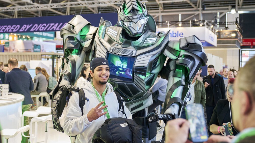 Person posing with a large greenish robot at BAU 2025 while someone takes a photo. Other visitors can be seen in the background.