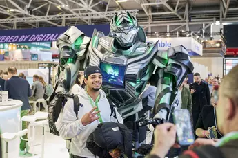 Person posing with a large greenish robot at BAU 2025 while someone takes a photo. Other visitors can be seen in the background.