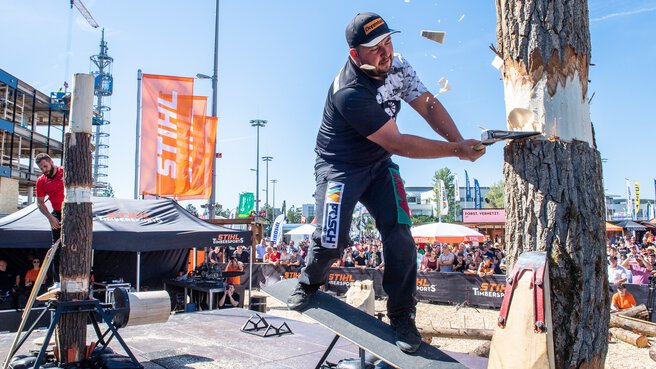 In a show area on the open-air grounds of INTERFORST, a man chops at an upright tree trunk with an axe in front of an audience.