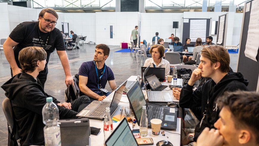 A group of people work with laptops at a free-standing table in a large exhibition hall.