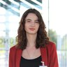 A young woman with curly brown hair wearing a red blazer stands in front of a bright, modern background.