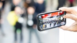 Eine Hand hält ein Smartphone, auf der die Kamerafunktion aktiviert ist. Auf dem Display ist eine Eingangshalle der Messe München mit zahlreichen Besuchern zu sehen.