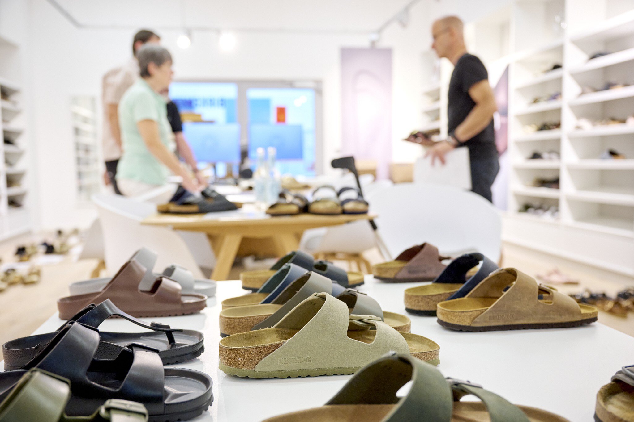 Eine Auswahl an bunten Sandalen steht auf einem Tisch. Im Hintergrund sind drei Personen in einem Ausstellungsraum zu erkennen.