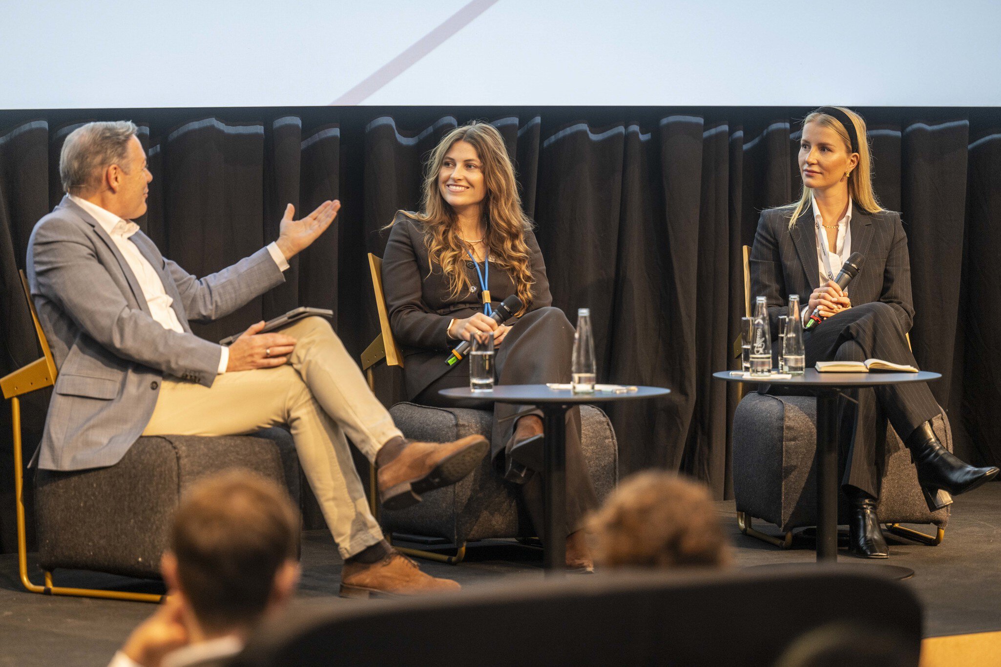 Three people are seated on stage in lounge chairs, engaged in a panel discussion. The man on the left is gesturing while the two women on the right listen attentively, each holding a microphone. Small tables in front of them hold water glasses and bottles.