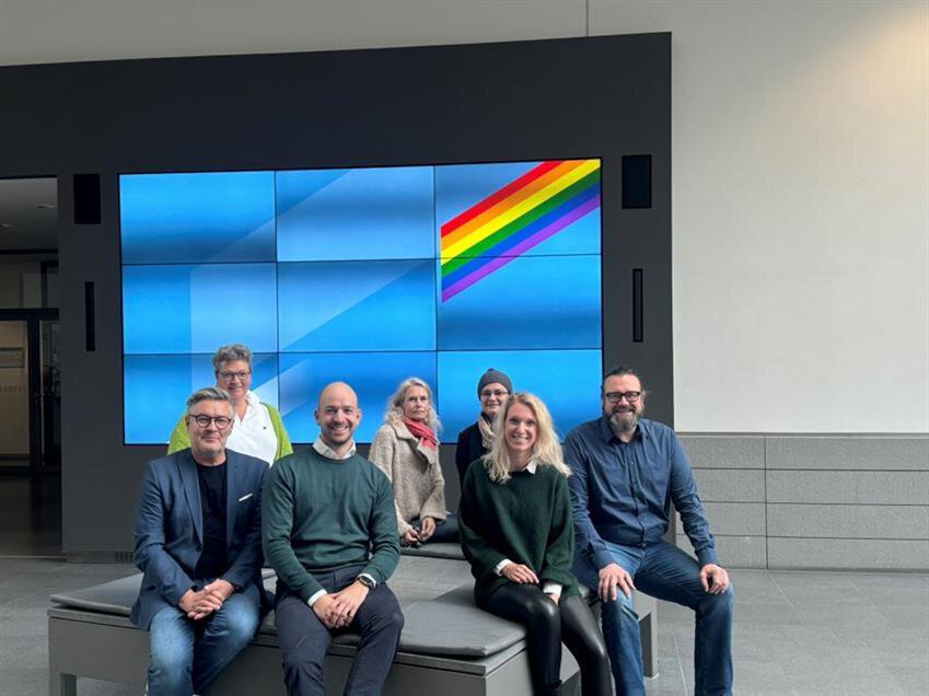 Eine Gruppe von sieben Personen sitzt gemeinsam vor einer blauen Monitorwand, in deren rechter oberer Ecke ein diagonaler Streifen mit Regenbogenfarben zu sehen ist.