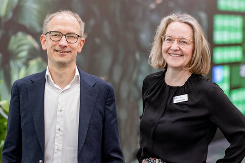 Zwei lächelnde Personen in Businesskleidung (Mann im dunkelblauen Sakko, Frau mit schwarzer Bluse und Namensschild) vor unscharfem, blättrigem Messestand mit Pflanzen.