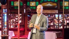 Ein Redner im grauen Anzug steht auf einer Bühne vor einem sitzenden Publikum, spricht mit Handgesten und hält einen Presenter in der Hand; im Hintergrund sind gedeckte Tische und bunt verglaste Fenster eines Veranstaltungssaals zu sehen.