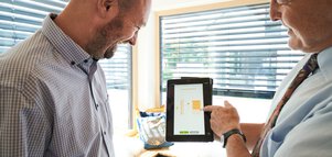 Ein Mann demonstriert einem anderen auf einem Tablet die Fernbedienung der Jalousien für das Fenster im Hintergrund.