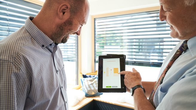 Ein Mann demonstriert einem anderen auf einem Tablet die Fernbedienung der Jalousien für das Fenster im Hintergrund.