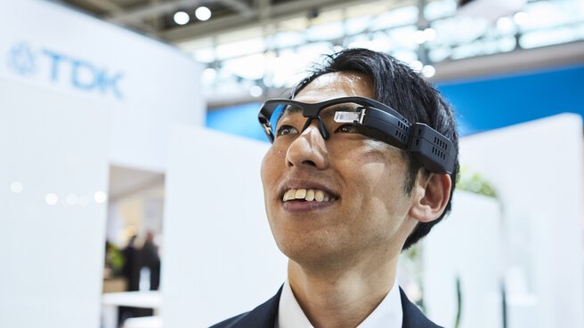 Lächelnde Person mit Datenbrille, im Hintergrund Messestände.