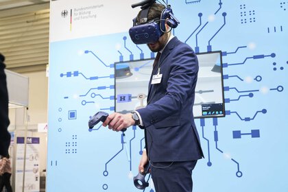 A person in a suit uses a VR headset and a controller at a technology stand with a digital display in the background.