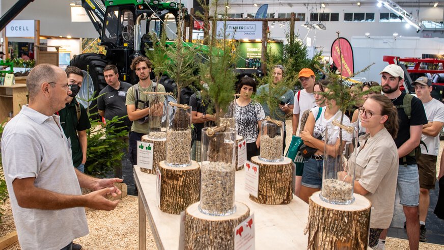 An exhibitor speaks to a group of young people standing around a table with tree seedlings piled up to varying heights with gravel.
