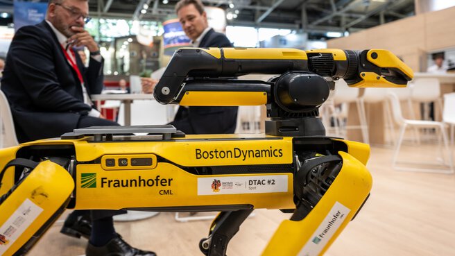 Two men in suits look at a yellow robot that moves like a dog and has a gripper arm. It was developed by the Fraunhofer Institute.