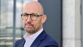 A man wearing glasses and a blue jacket stands with his arms crossed in front of a building with reflective glass windows.