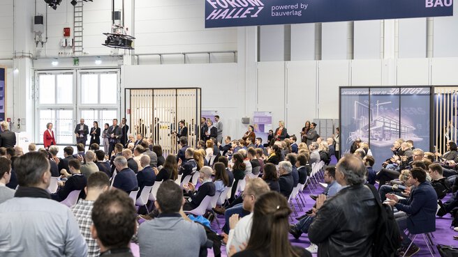Ein Publikum aus sitzenden und stehenden Menschen während einer Präsentation im Forum Halle 1 auf der digitalBAU.