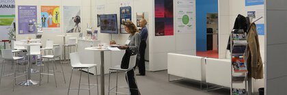 Trade fair visitors gathered at tables at an exclusive stand, surrounded by colorful posters and a screen adorning the walls.