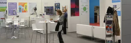 Messebesucher versammelten sich an Tischen eines exklusiven Standes, umgeben von bunten Plakaten und einem Bildschirm, der die Wände schmückte.