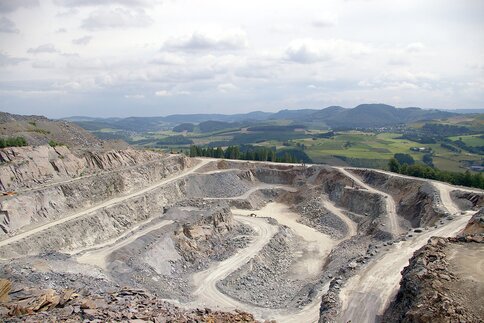 A large open-cast mine with terraced plains, surrounded by green hills and clouds in the sky.